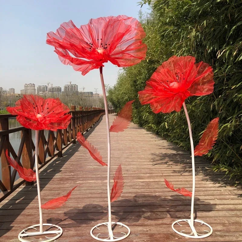 Three large red velvet mesh flowers on metal stems displayed along a wooden walkway next to green foliage.
