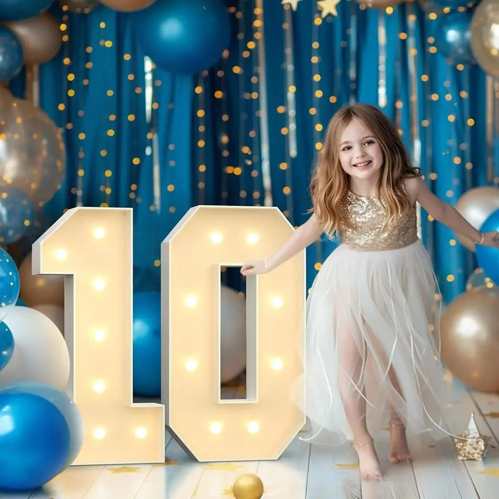 A young girl stands next to large light-up number balloons spelling '10' in front of a blue curtain backdrop and gold balloon
