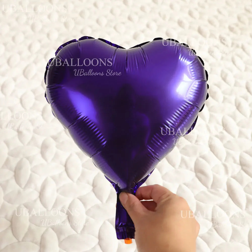 A purple heart-shaped foil balloon is being held by a hand against a white, textured background.