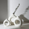 White ceramic vase with a donut pattern and a sprig of greenery on a white surface.