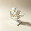 A white statue of a skeleton ghost reading a book is placed on a toilet bowl and stand.