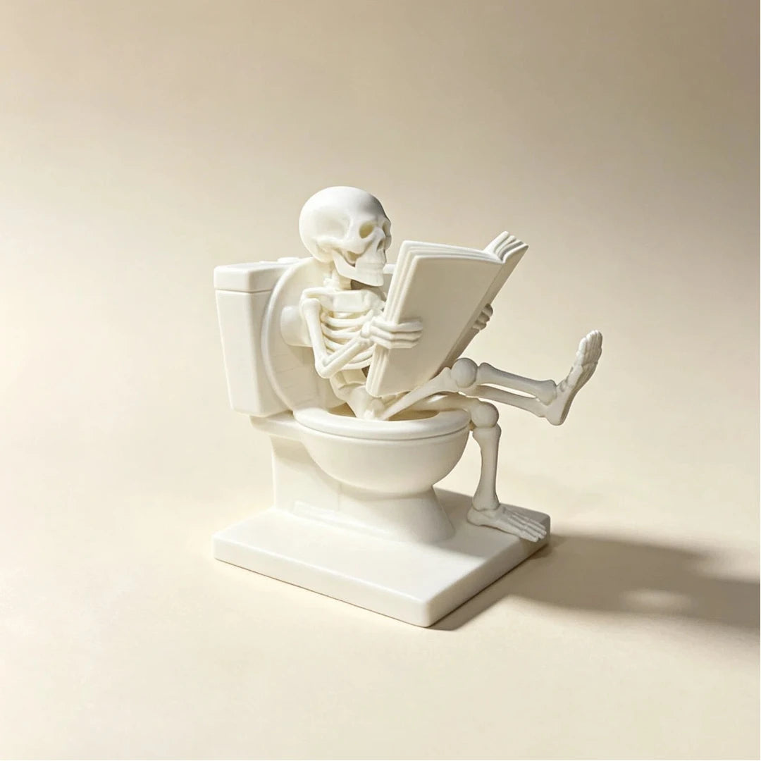 A white statue of a skeleton ghost reading a book is placed on a toilet bowl and stand.