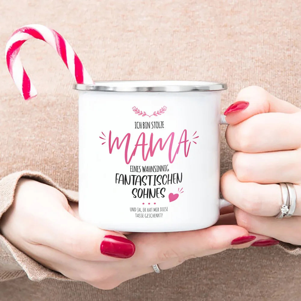 A white enamel mug with pink text reading ICH BIN STOLZE MAMA and a red and white striped candy cane is held by a person.