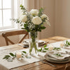 A clear glass vase filled with white roses and greenery on a wooden table.