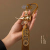A hand holds a brass gourd and five emperor coins hanging ornament made of gold-toned metal and silk thread.