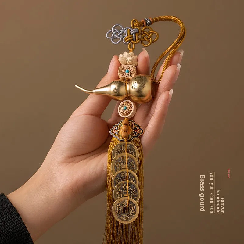 A hand holds a brass gourd and five emperor coins hanging ornament made of gold-toned metal and silk thread.