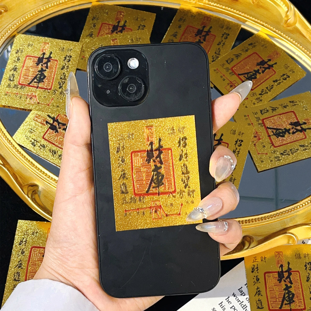A hand holds a black smartphone displaying a gold wealth amulet sticker against a background of yellow paper.