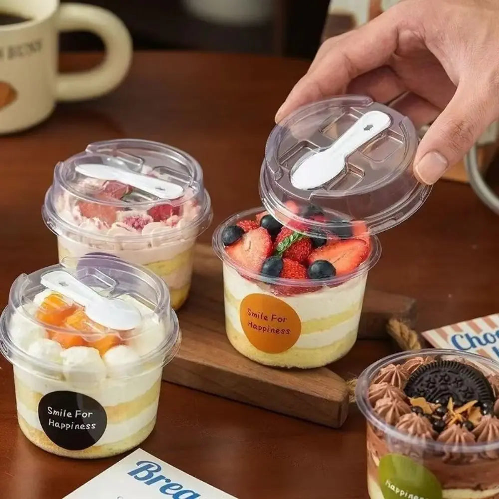 A hand holds a clear disposable pudding cup with a lid and spoon, resting on a wooden board.