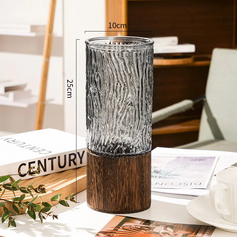 A tall, textured glass vase with a wooden base sits on a white surface.