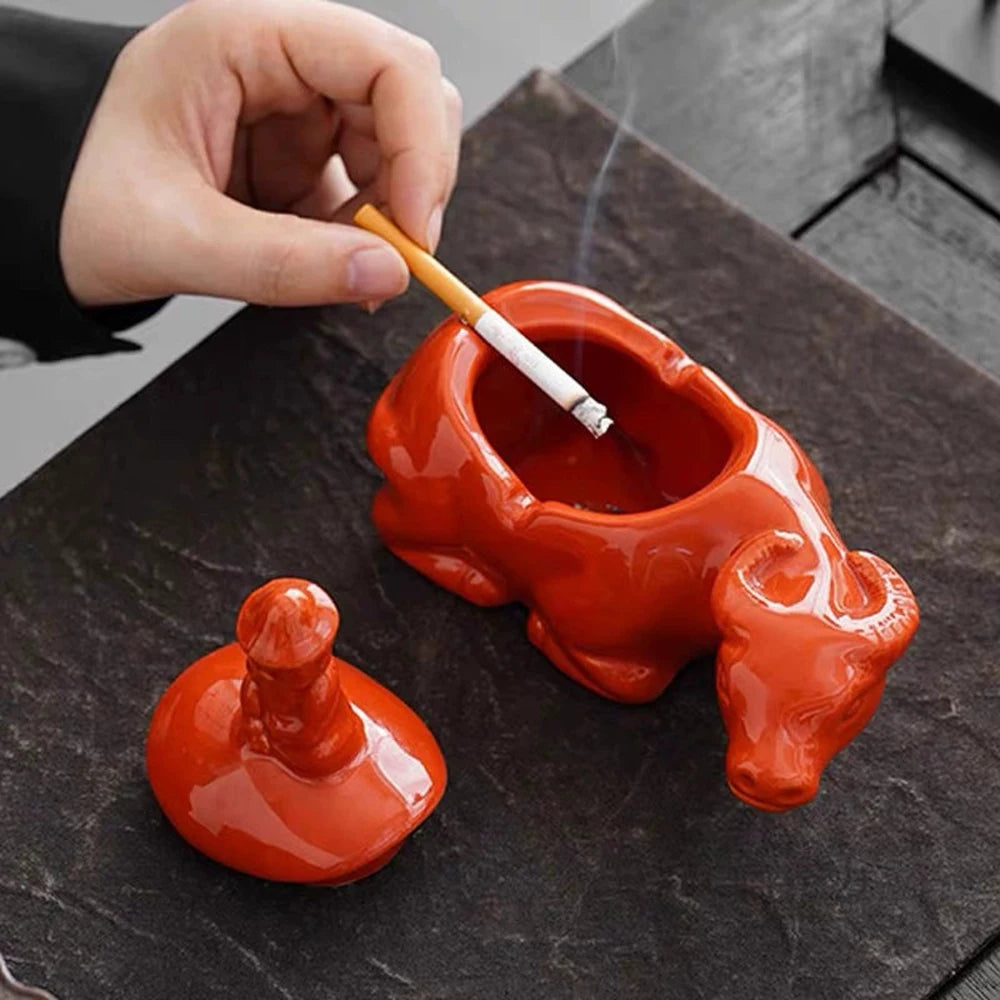 A hand holds a wooden stick over an orange ceramic crab ashtray with a lid on a dark slate surface.