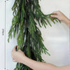 A hand is holding an artificial Norfolk pine garland with warm white lights against a white background.