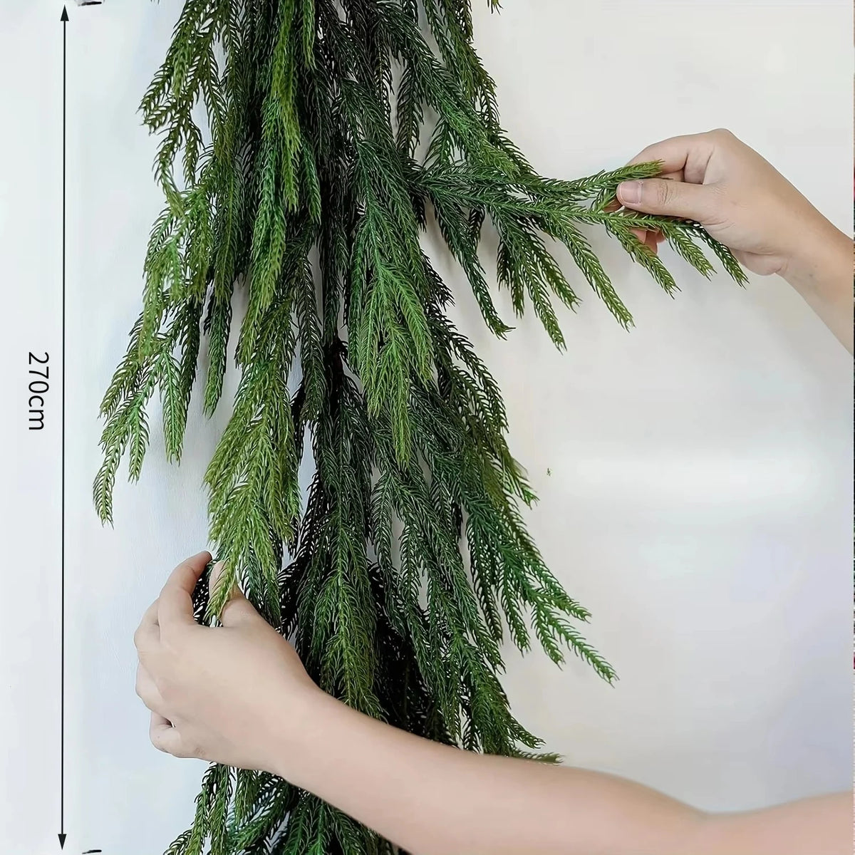 A hand is holding an artificial Norfolk pine garland with warm white lights against a white background.