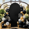 A dark velvet arch backdrop covered in black and gold balloons surrounded by green foliage.
