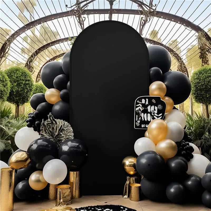 A dark velvet arch backdrop covered in black and gold balloons surrounded by green foliage.