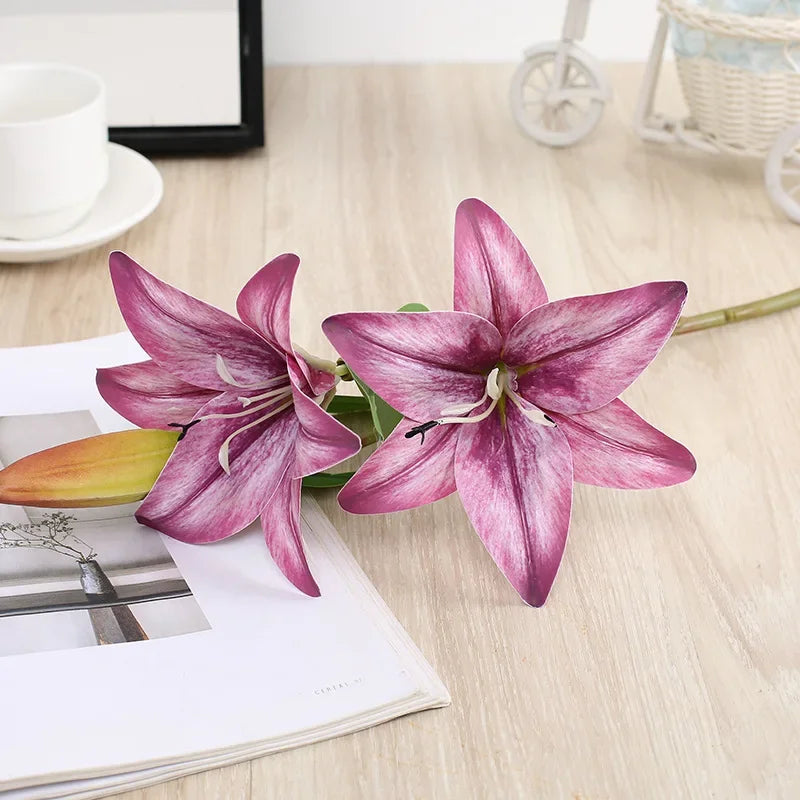 A realistic silk lily flower stem with three purple-pink blooms resting on a white paper.