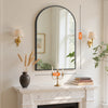 A bathroom setting featuring an arched mirror above a marble mantelpiece with decorative lamps and greenery.