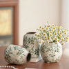 A white ceramic vase with green floral patterns sits on a wooden surface next to a pearl necklace.