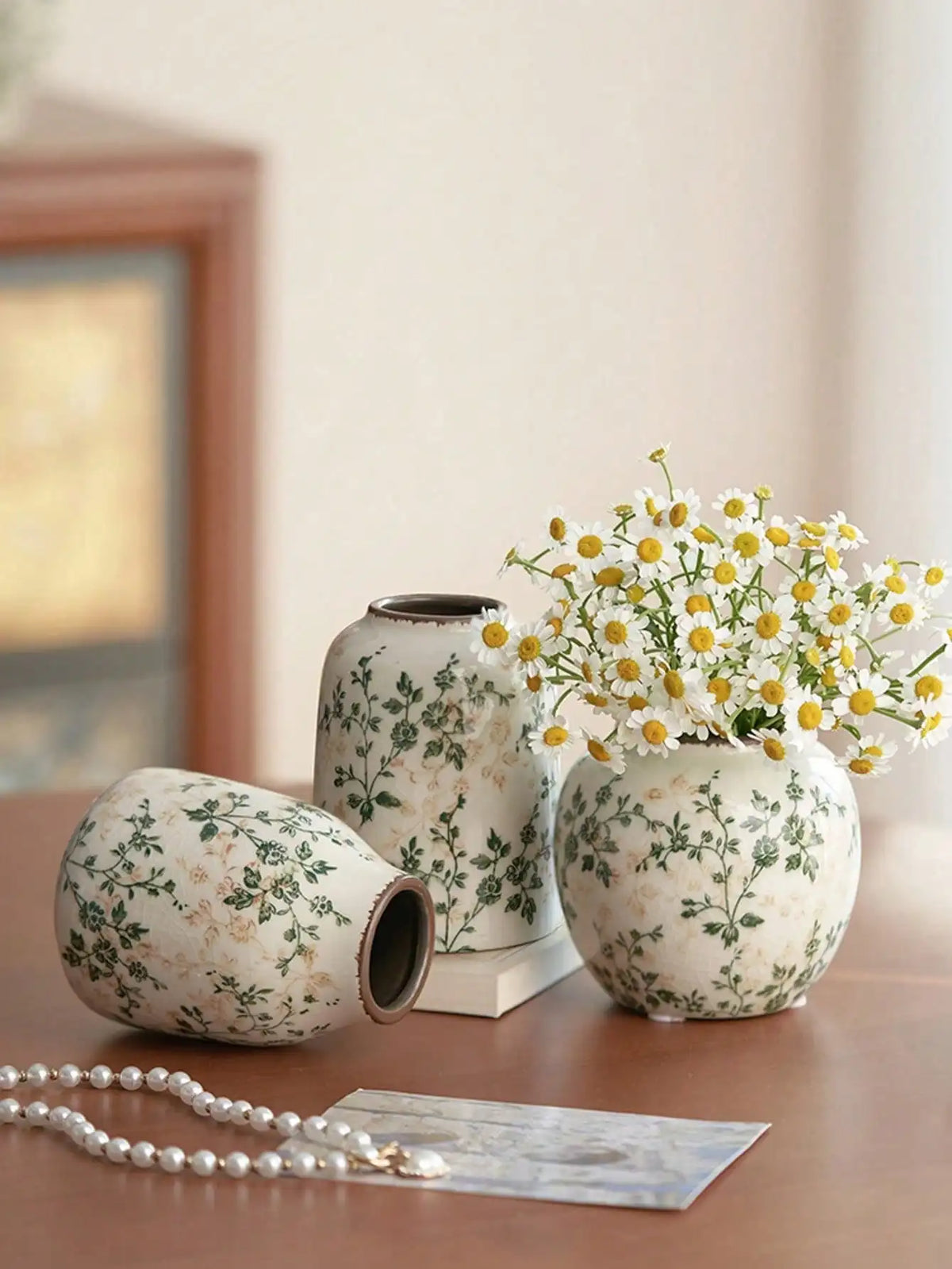 A white ceramic vase with green floral patterns sits on a wooden surface next to a pearl necklace.