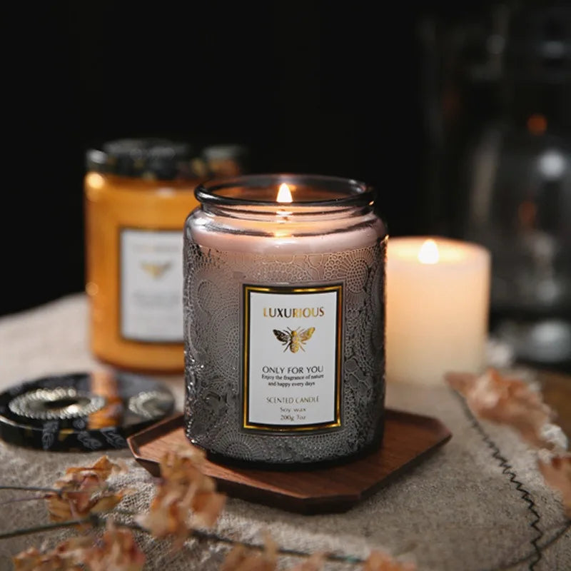 A jar of luxurious scented soy candle sits on a wooden coaster with dried botanicals in the background.