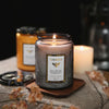 A jar of luxurious scented soy candle sits on a wooden coaster with dried botanicals in the background.