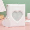 A white candy heart window photocard album with a silver chain attached, resting on a pink surface.