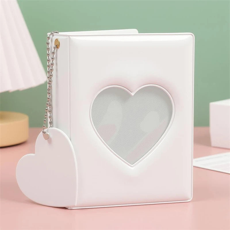 A white candy heart window photocard album with a silver chain attached, resting on a pink surface.