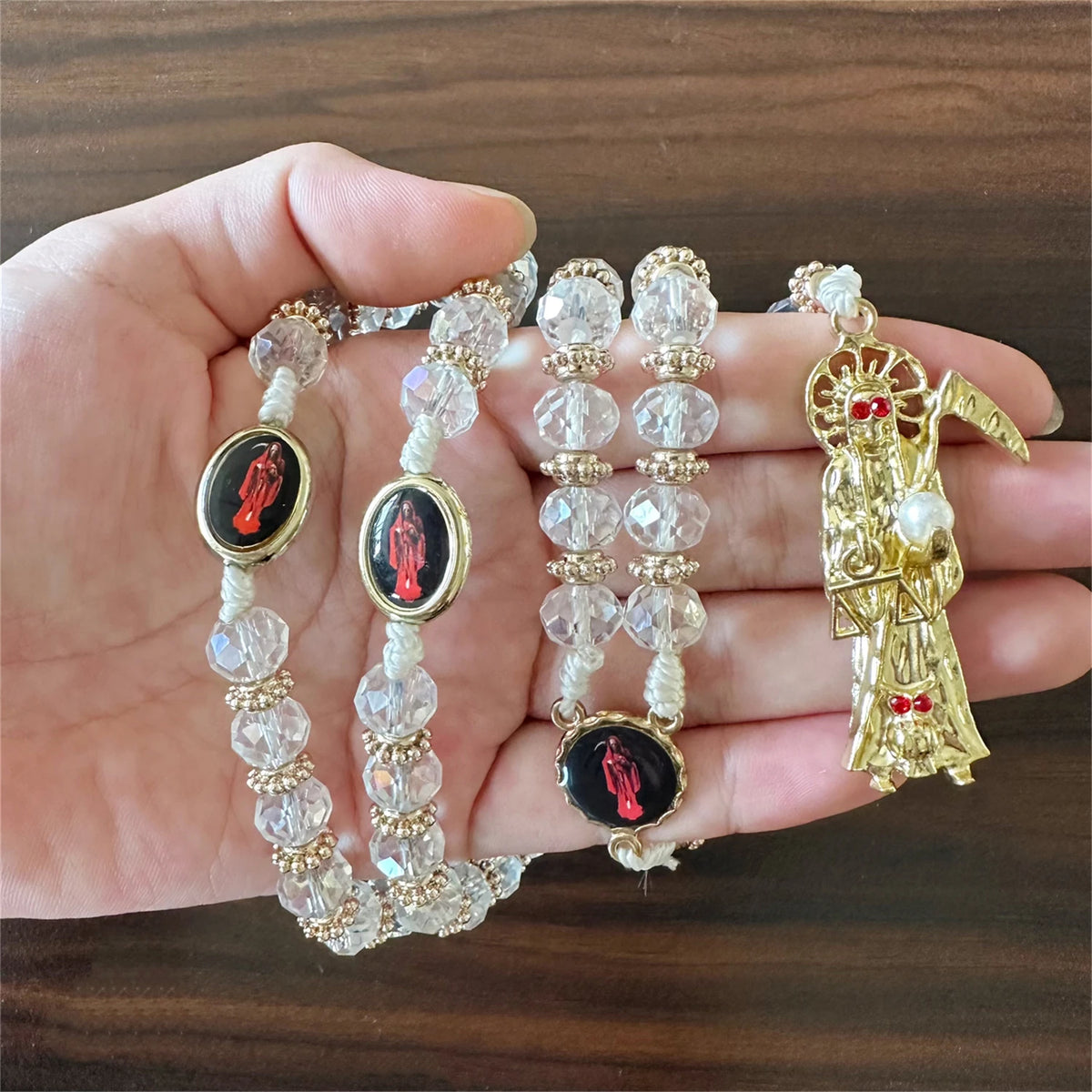 A hand holds a rosary necklace featuring clear beads and black oval medallions with red figures, alongside a gold Santa Muert