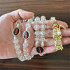 A hand holds a rosary necklace featuring clear beads and black oval medallions with red figures, alongside a gold Santa Muert