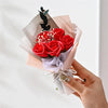 A hand holds a bouquet of red roses crafted from handcrafted soap, wrapped in pink and gray paper.