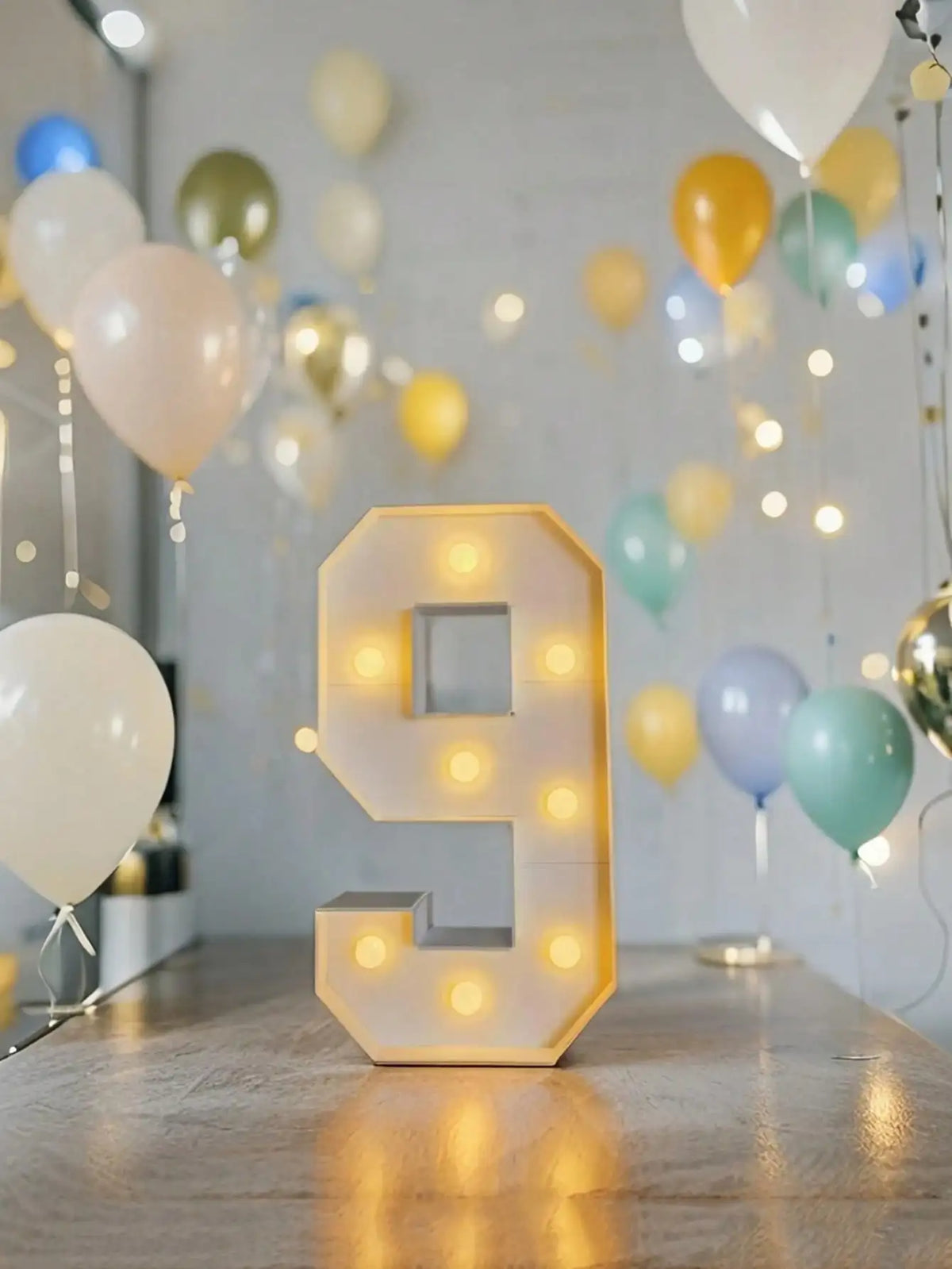 A glowing yellow LED marquee number 9 in a square frame sits on a wooden surface amidst balloons and string lights.