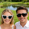 A smiling bride and groom wearing pearl decor glasses for wedding photos outdoors.
