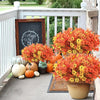 A vibrant display of artificial orange and yellow flowers in a pot sits on a porch railing next to pumpkins.