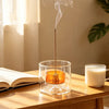 A gourd-shaped glass incense burner with an orange-colored stick is smoking on a wooden table.