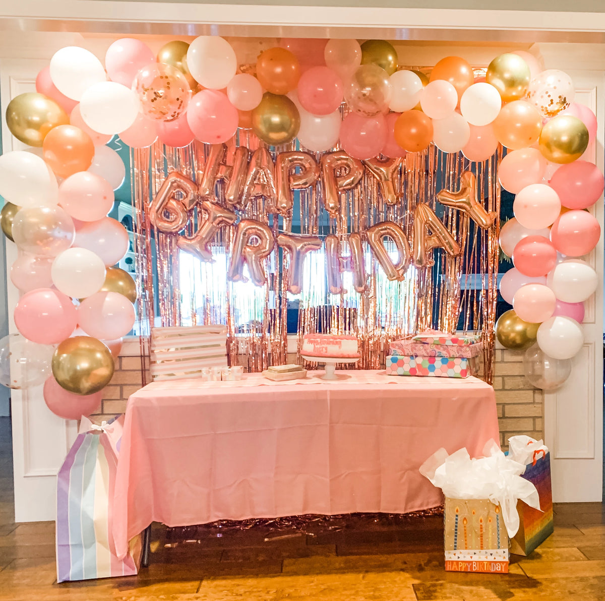 A pink and gold balloon arch backdrop decorated with metallic tinsel foil fringe curtains for a birthday party.