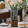A tall glass vase with a wooden base holding green stems and pink roses.