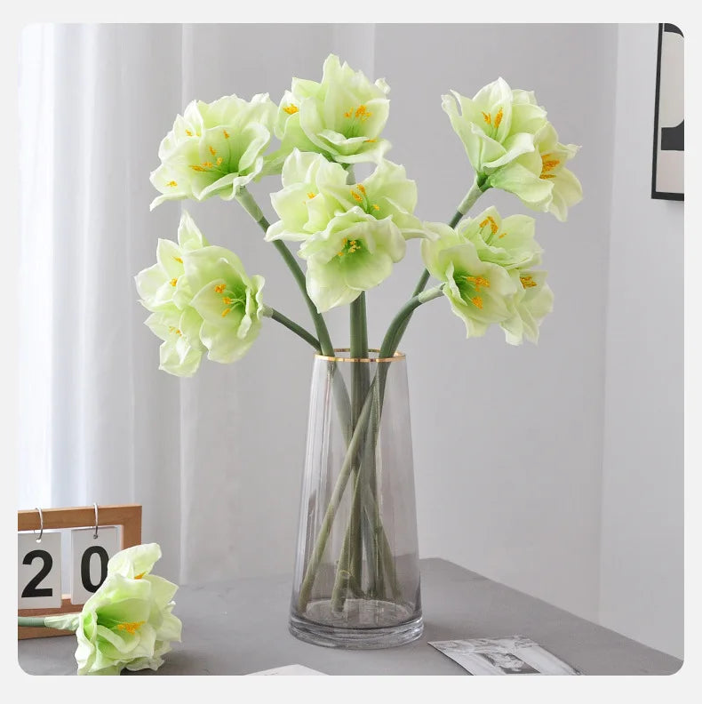 A vase holds several light green artificial clivia amaryllis flowers on a gray surface.