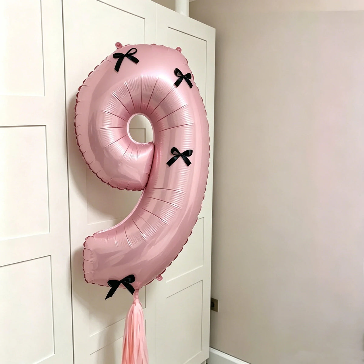 A pink foil balloon shaped like the number 9 with black bows, hanging on a white door.