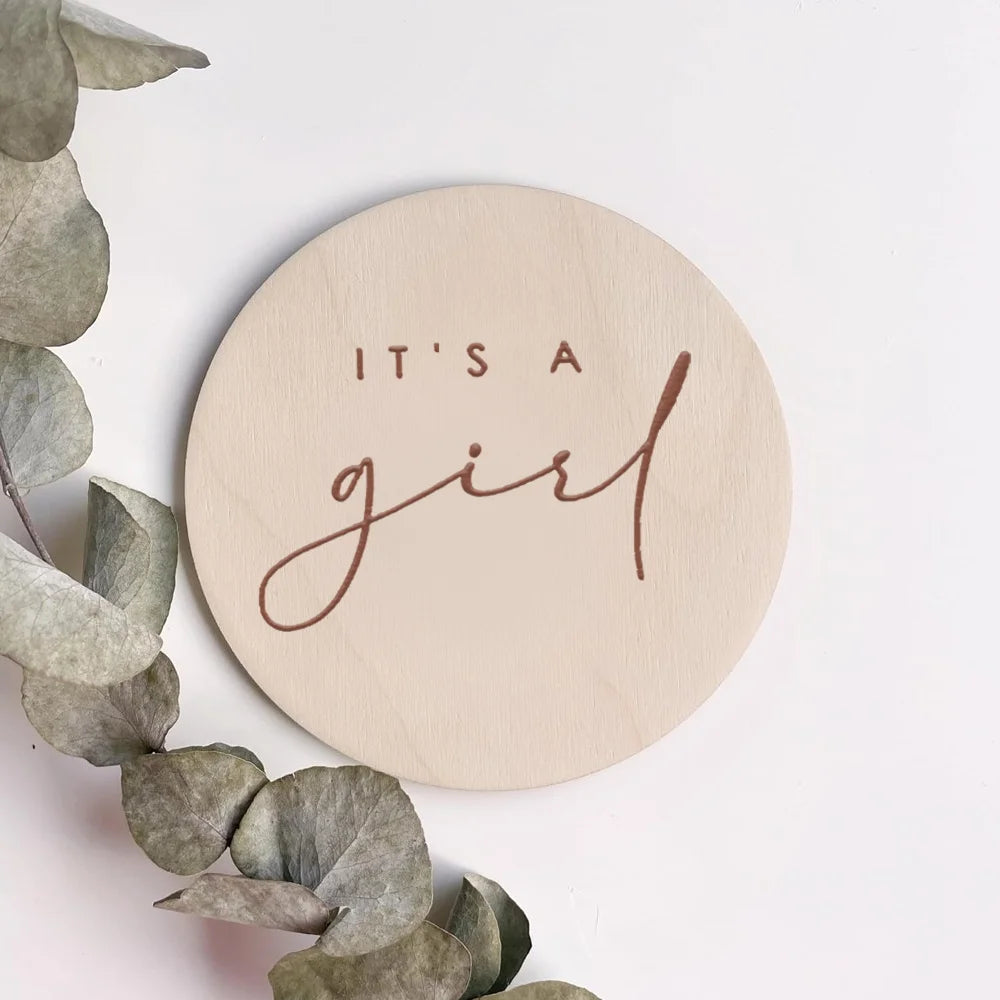 A round light wood plaque with the cursive text It's a girl engraved on it, surrounded by eucalyptus leaves.