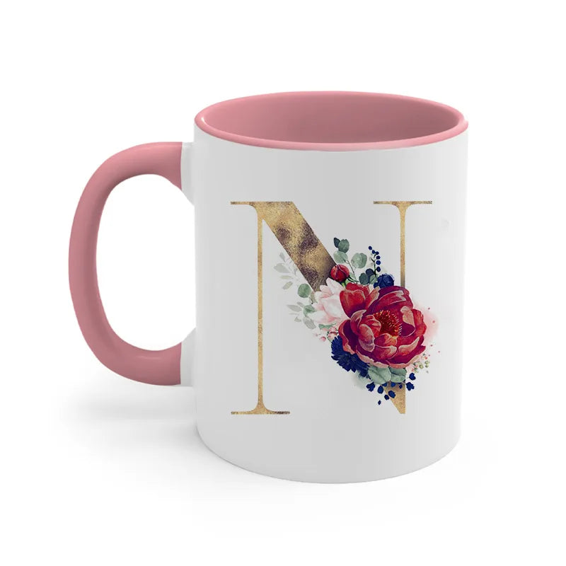 A pink ceramic coffee mug with a gold letter initial decorated with floral elements.
