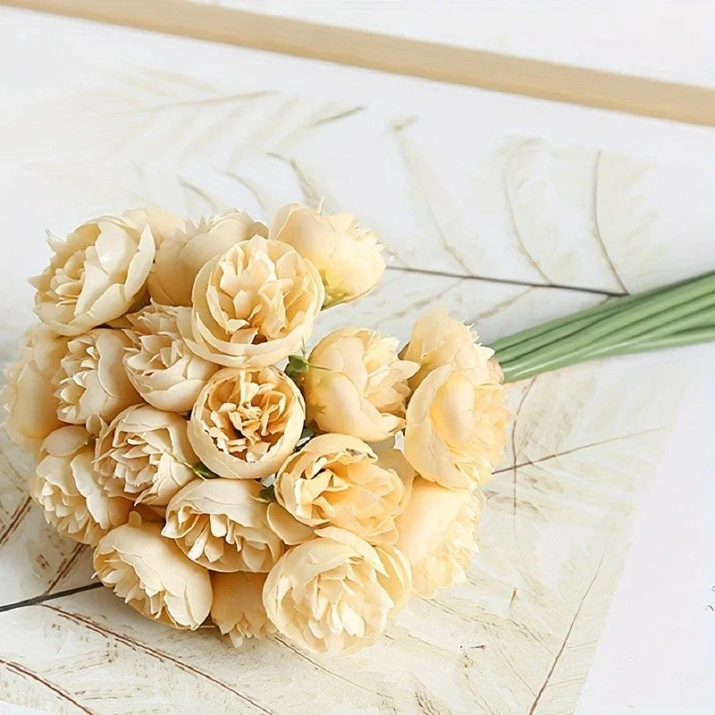 A bouquet of light yellow silk peonies with green stems arranged on a white surface.