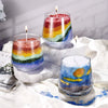 Three glass candles in rainbow colors are displayed on a white cloth with natural light.
