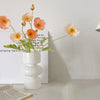 A white ceramic vase holds several delicate, handcrafted glass flowers with orange and pink petals.