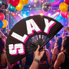 A hand holding a large black foldable clapper fan with white and pink letters spelling SLAY against a colorful party backgrou