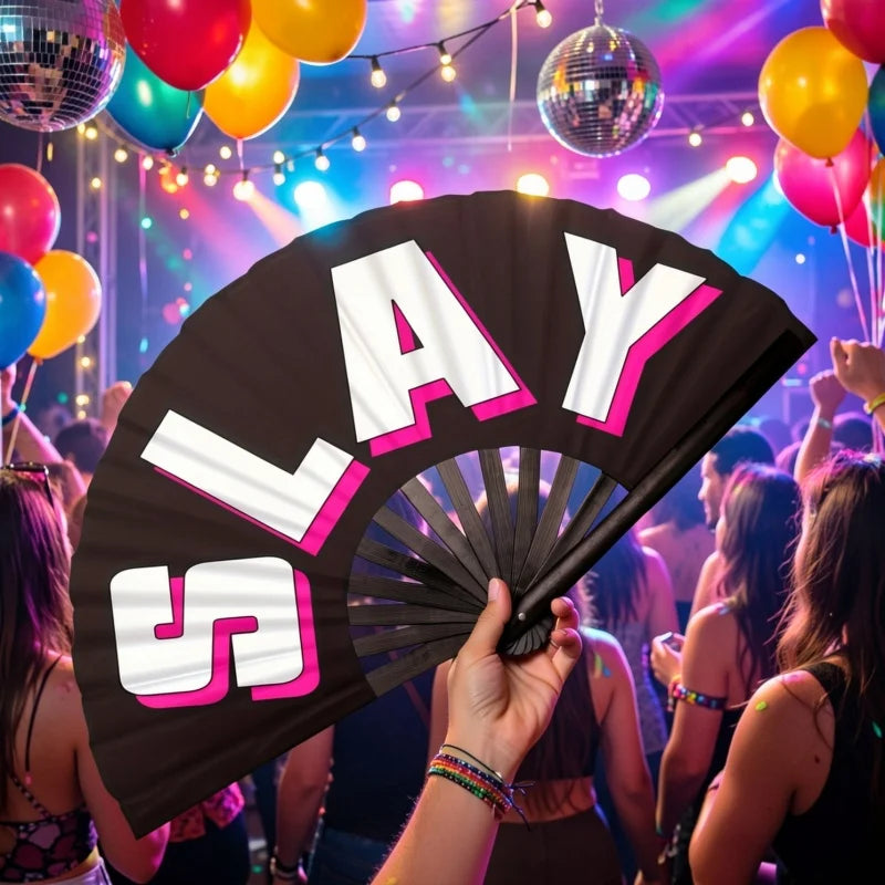 A hand holding a large black foldable clapper fan with white and pink letters spelling SLAY against a colorful party backgrou