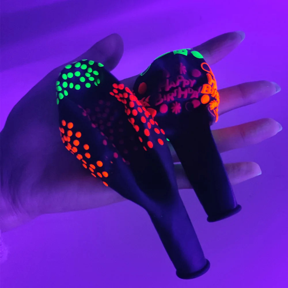 A hand holds a black latex balloon with neon green and orange glow-in-the-dark patterns against a purple background.