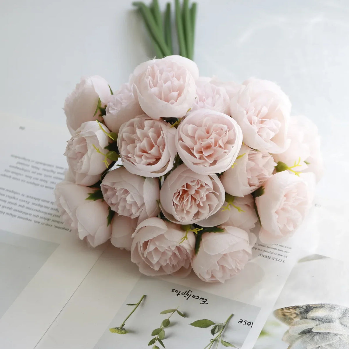A bouquet of artificial pink peony silk flowers arranged on white paper with green stems.