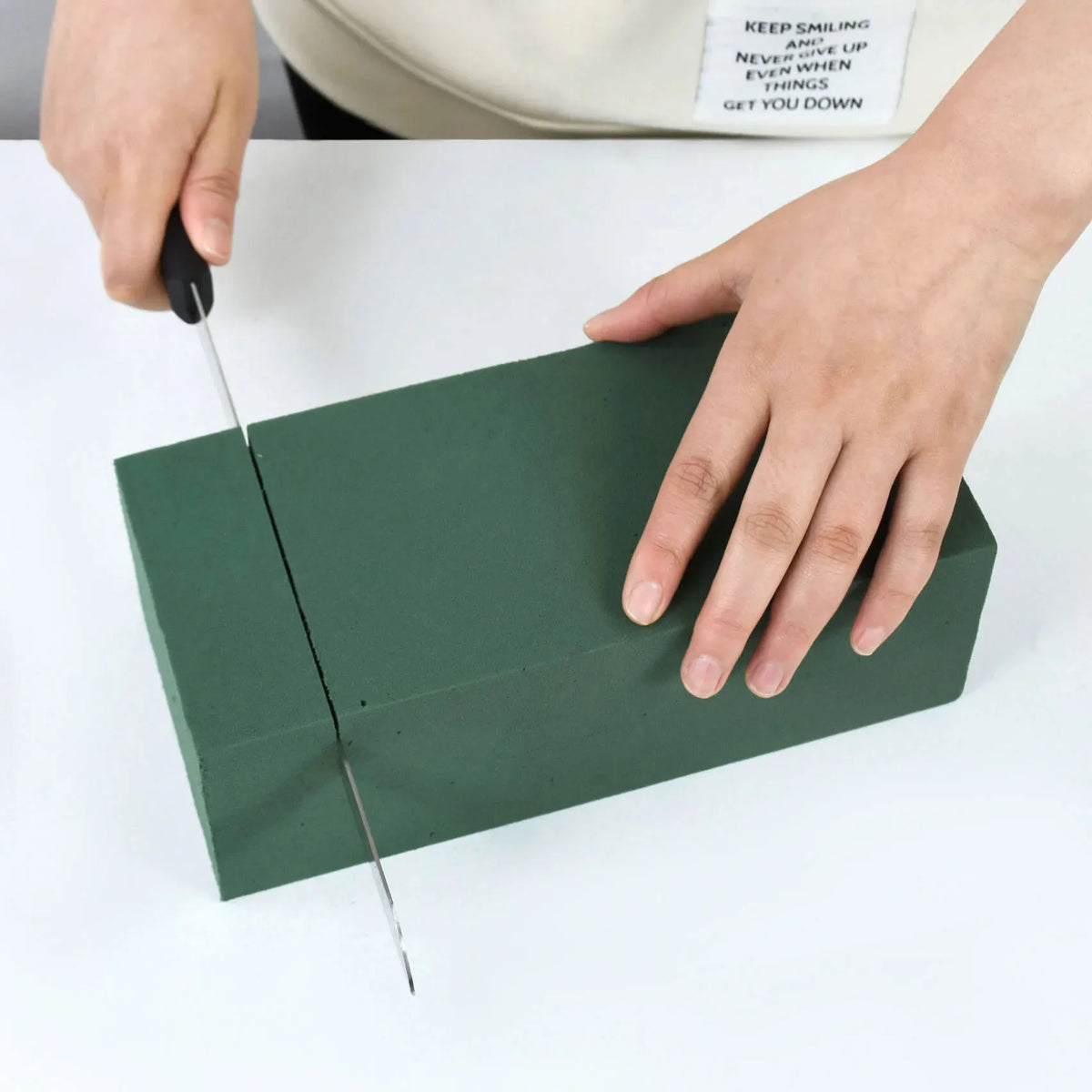 A hand is cutting a green floral foam block for party decor on a white surface.