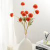 A vase holds a spray of bright orange artificial chrysanthemum pompon flowers on a white surface.