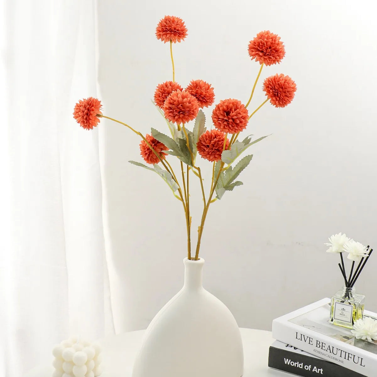 A vase holds a spray of bright orange artificial chrysanthemum pompon flowers on a white surface.