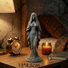 A gray praying angel figurine statue displayed on a wooden table next to a vintage gold alarm clock and a lit candle.